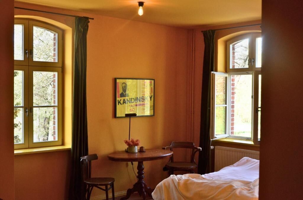 A room with two windows, a bed, a table with a vase, and a framed picture on the wall.