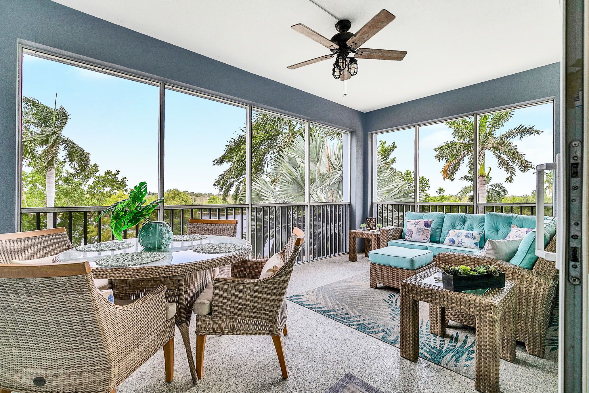 A sunroom with wicker furniture, a ceiling fan, and large windows offering a view of palm trees.