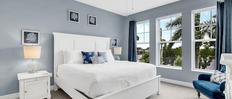 A bedroom with a white bed, a nightstand, a blue chair, a ceiling fan, and a window with curtains.