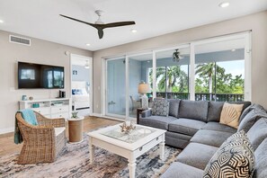 A spacious living room with a large grey sofa, a white coffee table, and a television. There is a balcony with a view of palm trees. - One Particular Harbour Lagoon View 391-203 · Pet-Friendly (Bradenton)