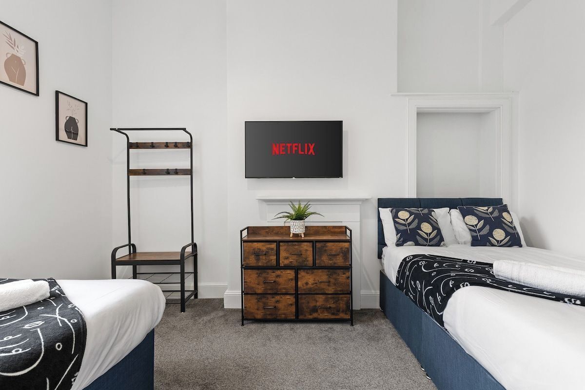 Un dormitorio con dos camas, un televisor de pared mostrando 'NETFLIX', un aparador de madera y una estantería metálica.