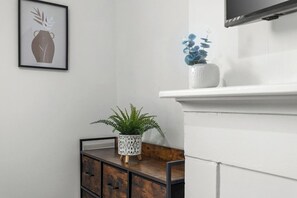 Una habitación con pared blanca, una obra de arte enmarco, un pequeño armario de madera con cajones, un helecho en maceta y un jarrón blanco con flores azules.