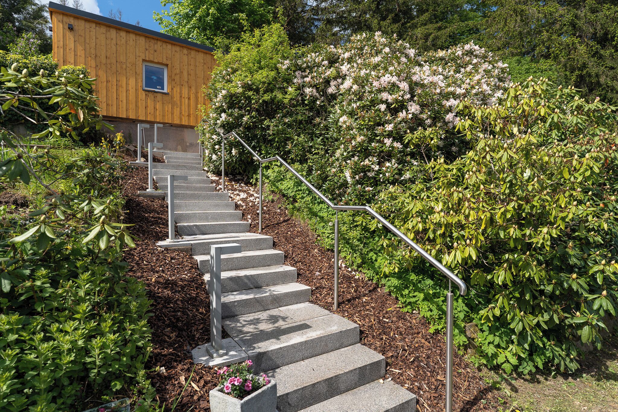 Eine Treppe aus Steinen führt nach oben zu einem Holzhaus, umgeben von üppigem Grün und blühenden Büschen.