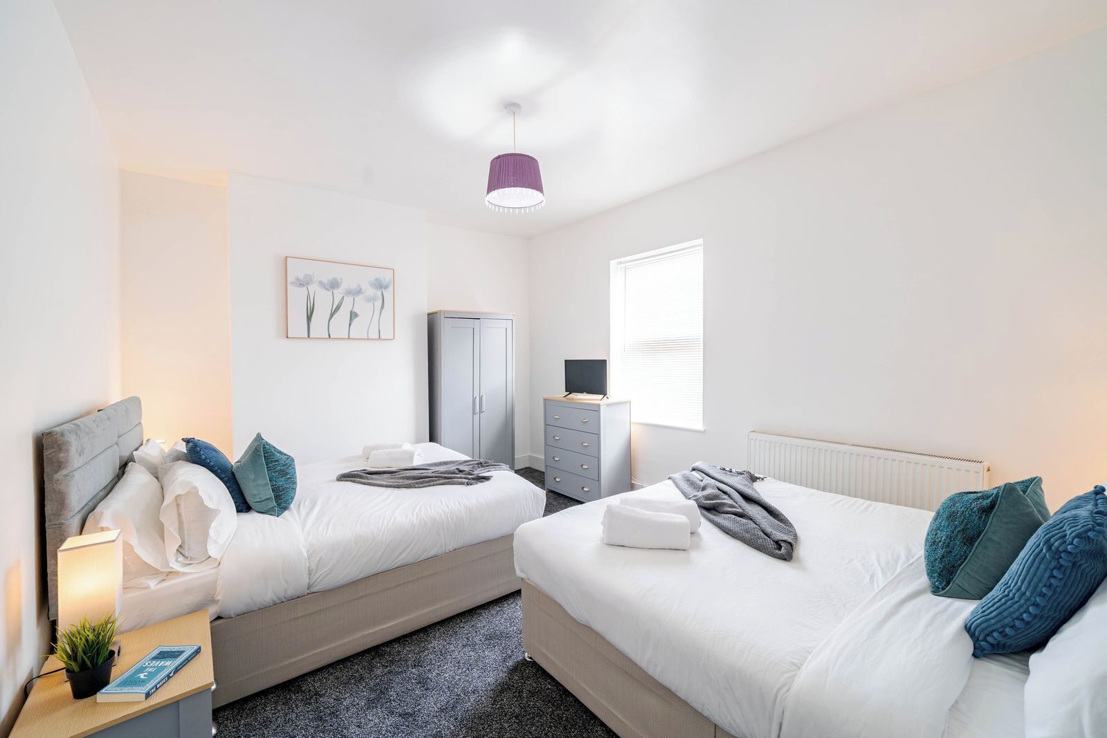 A hotel room with two beds, a wardrobe, a desk with a lamp, and a window with blinds.