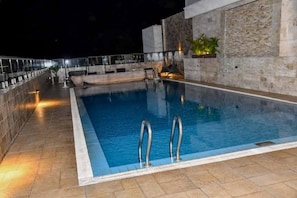 Une piscine rectangulaire avec deux échelles à la partie peu profonde, entourée d’une terrasse carrelée et d’un mur en pierre.