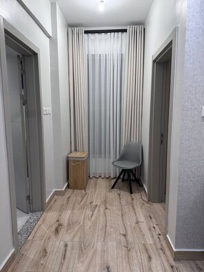 Ein Flur mit Holzboden, einem grauen Stuhl, einem kleinen Hocker und einem Fenster mit durchsichtigen Vorhängen.