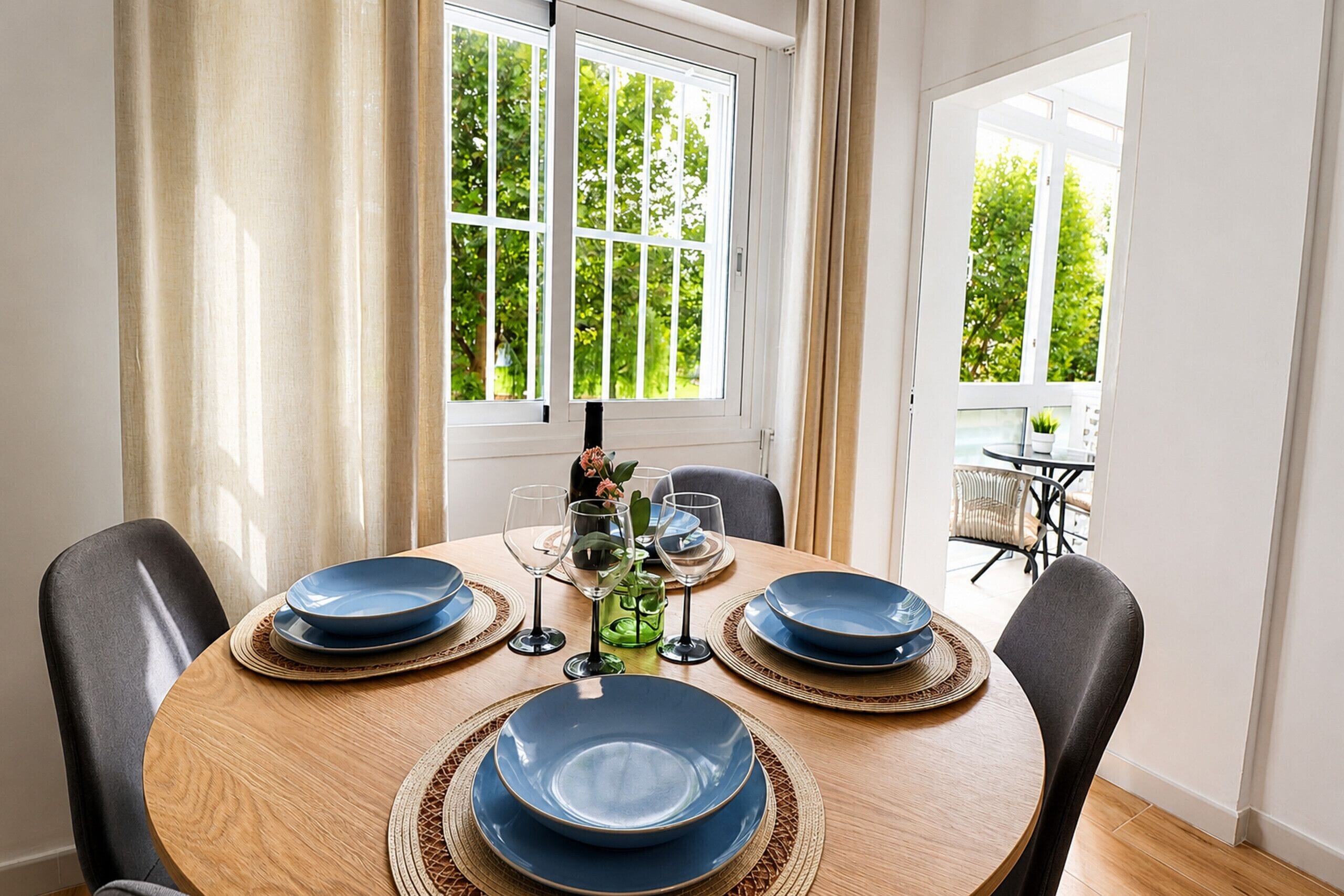A dining table with blue plates, wine glasses, and a vase with flowers, set against a window with a view of greenery.