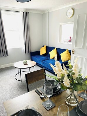 Um quarto com um sofá azul, uma mesa redonda e um espaço de refeições com pratos, copos e talheres.