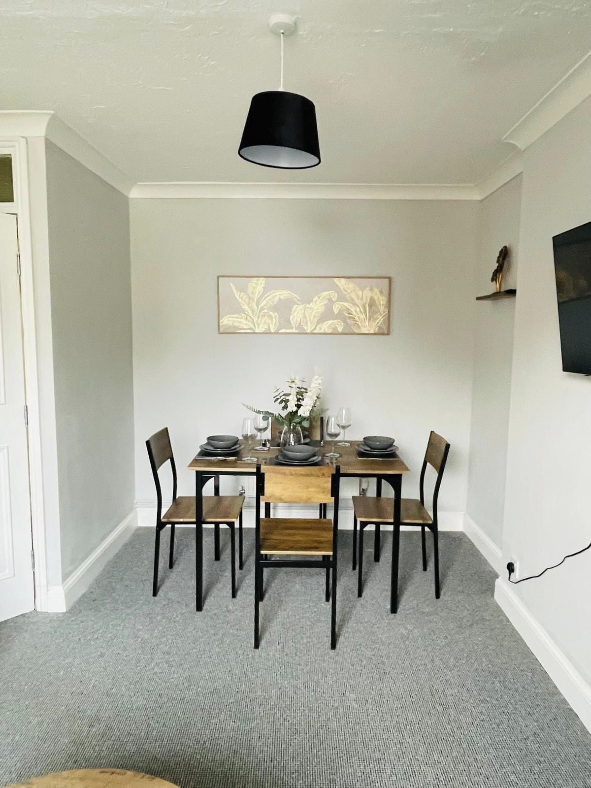 A dining area with a table set for four, a black pendant light, and a framed botanical print on the wall.