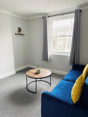 Um quarto com um sofá azul, uma pequena mesa redonda com uma planta, uma janela com cortinas e uma prateleira fixa na parede com um objeto decorativo.