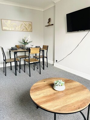 Ein Essbereich mit einem runden Holztisch, vier Stühlen, einer Blumenvase und einem an der Wand befestigten Flachbildfernseher.
