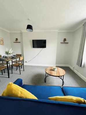 A living room with a blue sofa, a wooden coffee table, a dining table with chairs, a flat-screen TV mounted on the wall, and a small plant on a shelf. - Seaside Escape Lowestoft | Stylish Coastal Stay (Kirkley)