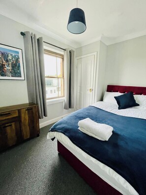 Um quarto com cama, janela, porta, mesa de cabeceira e luminária suspensa.
