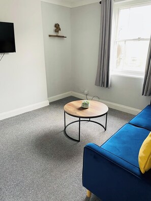 Ein Wohnzimmer mit einem blauen Sofa, einem runden Couchtisch aus Holz, einem grauen Teppich, einem Wand-Fernseher, einer kleinen Ablage mit einer Dekoration und einem Fenster mit Vorhängen.
