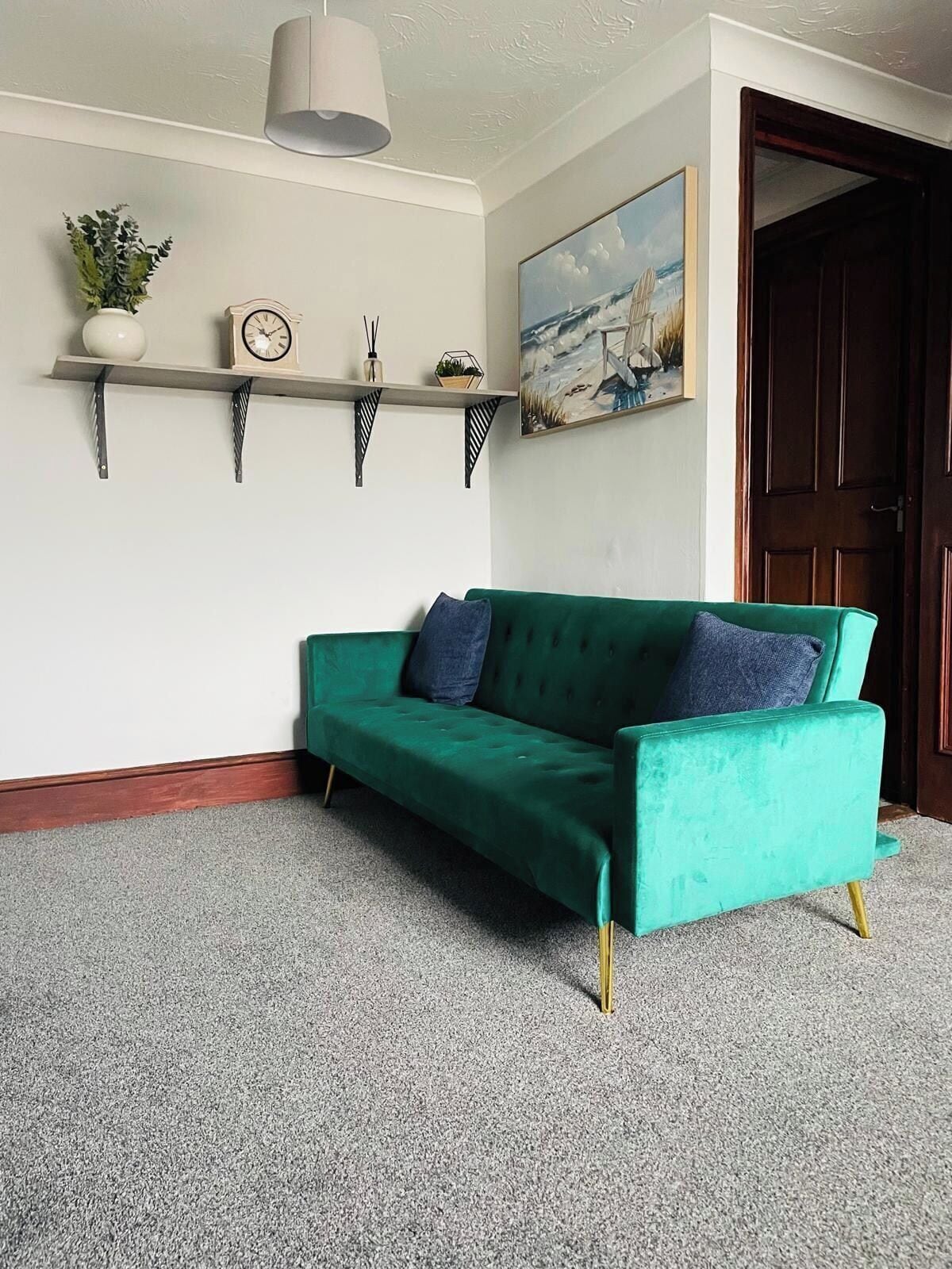 A green sofa with gold legs, a white wall, a clock, a plant, and a painting.