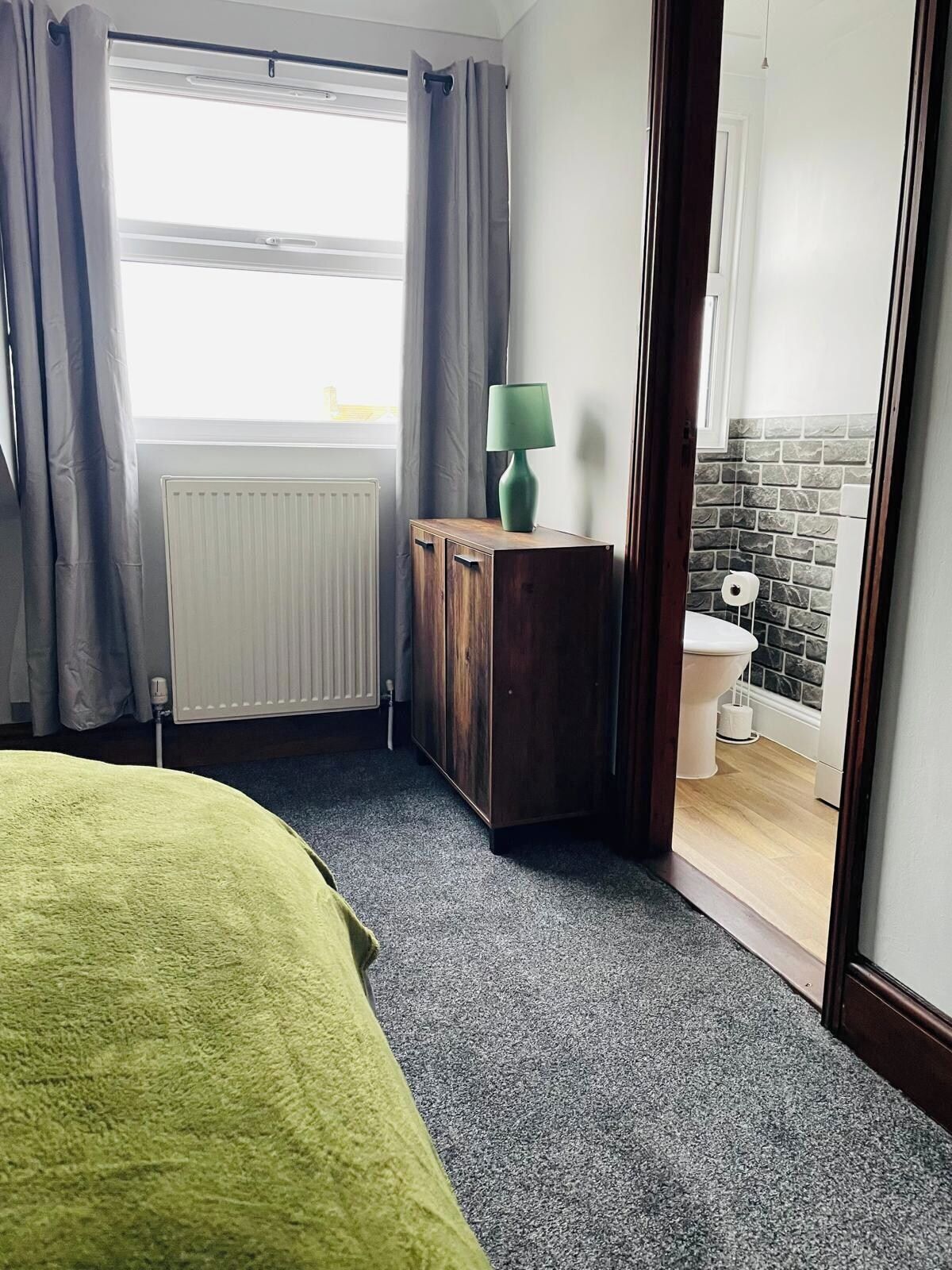 A bedroom with a bed, a radiator, a wooden cabinet, a green lamp, and a view of a bathroom with a toilet and a brick wall.