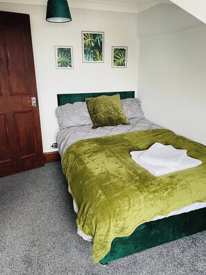 Un dormitorio con cama, cabecera verde, ropa de cama gris, una manta verde, una toalla blanca y tres cuadros con hojas tropicales en la pared.