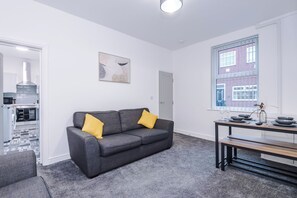 A modern living room with a grey sofa, a dining table with chairs, a window with a view of a brick building, and a kitchen in the background. - Spacious Home with Free Parking | Atherton House (Greater Manchester)