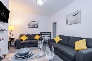 A modern living room with two grey sofas, yellow cushions, a dining table with plates and cutlery, a wine glass, a lamp, and artwork on the walls. - Spacious Home with Free Parking | Atherton House (Greater Manchester)