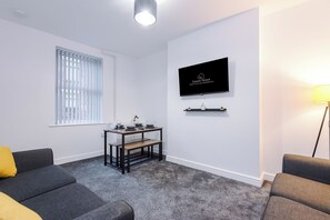 Een moderne woonkamer met een grijze bank, een eettafel met een bankje, een aan de muur bevestigde televisie en een vloerlamp.