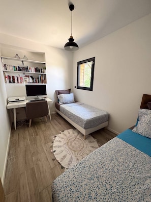 Ein Schlafzimmer mit einem Bett, einem Schreibtisch mit Computer, einem Bücherregal und einem Fenster.