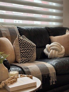 A dark leather sofa with a blanket and multiple cushions, a small table with a book and candles, and a window with blinds. - Spacious Stay by the Docks | Your Wallasey Getaway (Liverpool / Merseyside)