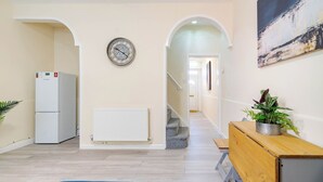 A hallway with a white radiator, a clock, a refrigerator, a bench, and a staircase leading to another room. - Rishton Stay | Shops & Transport | Near Blackburn (Lancashire)