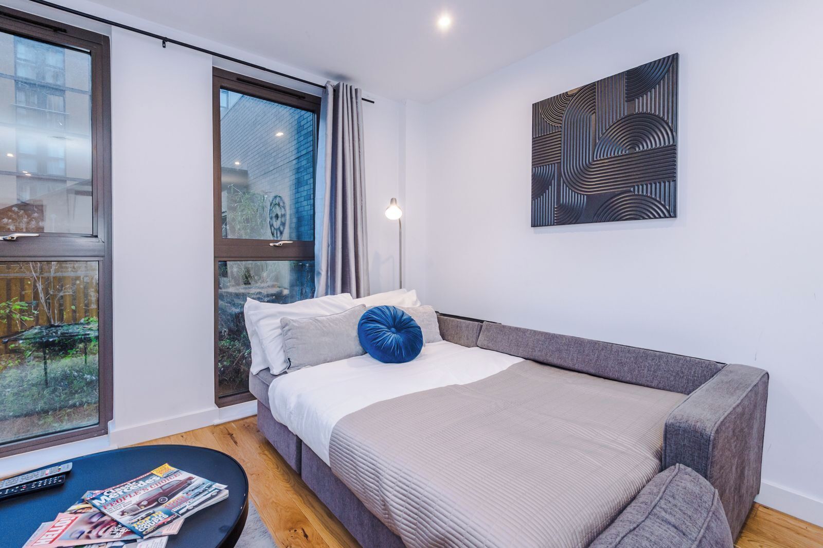 A modern bedroom with a large window, a bed with a grey headboard, and a wall-mounted abstract art piece.