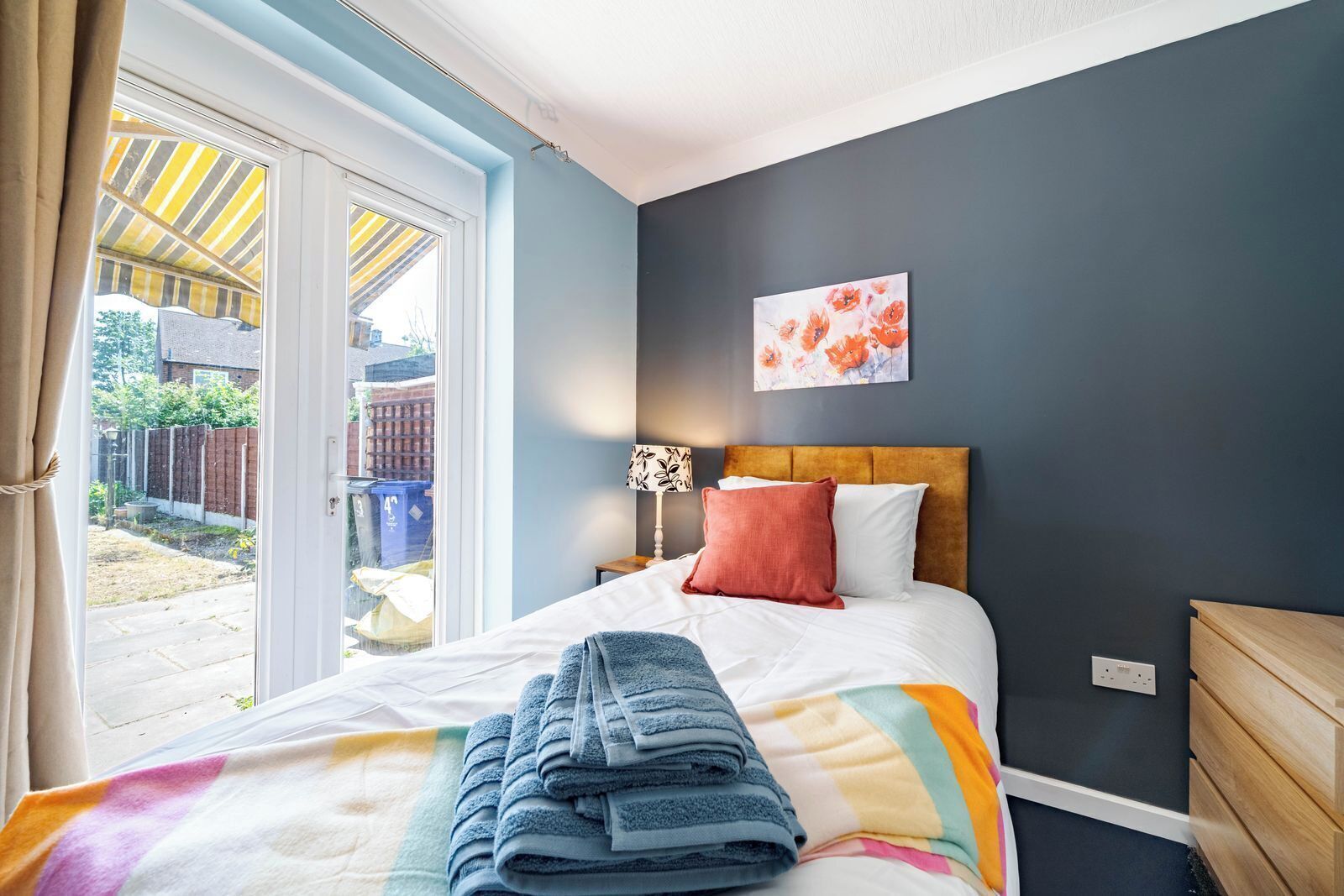 A bedroom with a bed, a nightstand, a lamp, a wall art piece, and a view of the outside.