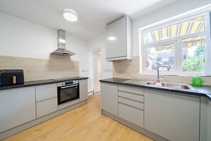 A modern kitchen with a built-in oven, a stainless steel range hood, and a window with striped curtains. - Wythenshawe Stay | Airport & Hospital | Parking (Greater Manchester)