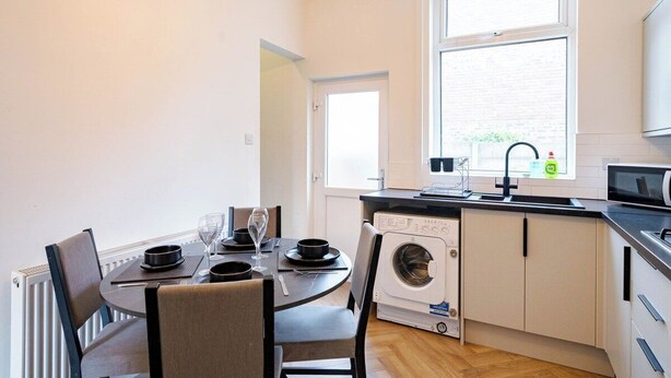 Une cuisine compacte équipée d’un lave-linge, d’un coin repas avec une table et des chaises, et d’une fenêtre laissant entrer la lumière naturelle.