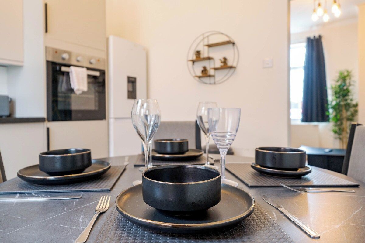 A modern kitchen with a dining table set for two, featuring black plates, bowls, and cutlery.