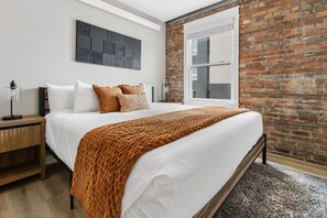 A bedroom with a brick wall, a large bed with white bedding, a brown throw blanket, and a modern art piece above the bed. - Historic Brownstone Apartment, Heirloom Vacations (Duluth)