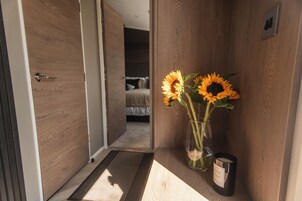 Un pasillo con una puerta de madera, una cama al fondo y un florero con girasoles sobre una estantería.