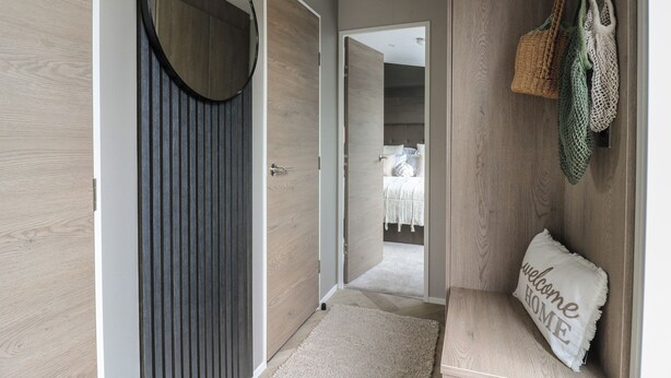 A hallway with a mirror, a door, a bench with a pillow, and a wall with hanging bags.