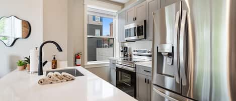 A modern kitchen with stainless steel appliances, a large island with a sink, and a window overlooking the street.