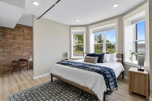 A bedroom with a large bed, a wooden nightstand, a chair, and a view of the outdoors through large windows. - Historic Brownstone Apartment, Heirloom Vacations (Duluth)