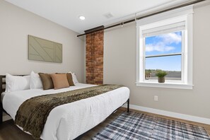 Un dormitorio con cama, ventana con vistas, pared de ladrillo y una lámpara de techo.