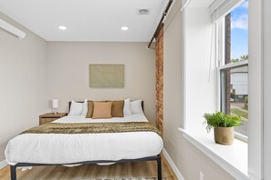 Ein Schlafzimmer mit Bett, Nachttisch, Fenster und Blick sowie einer Pflanze auf der Fensterbank.