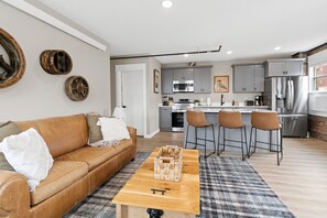 Ein modernes Wohnzimmer mit einem braunen Ledersofa, einem Holztisch und einer Küche im Hintergrund, die graue Schränke und Edelstahlgeräte zeigt.