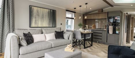 A modern living room with a grey sofa, a coffee table, and a black armchair. There is a dining area with a table and chairs, and a kitchen with a fridge and stove.