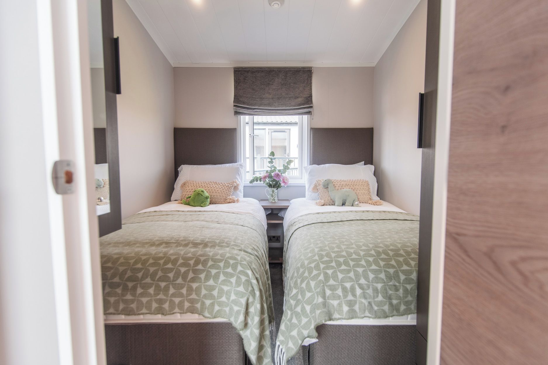 Una habitación estrecha con dos camas, una ventana con cortinas y una pequeña mesa con un florero.