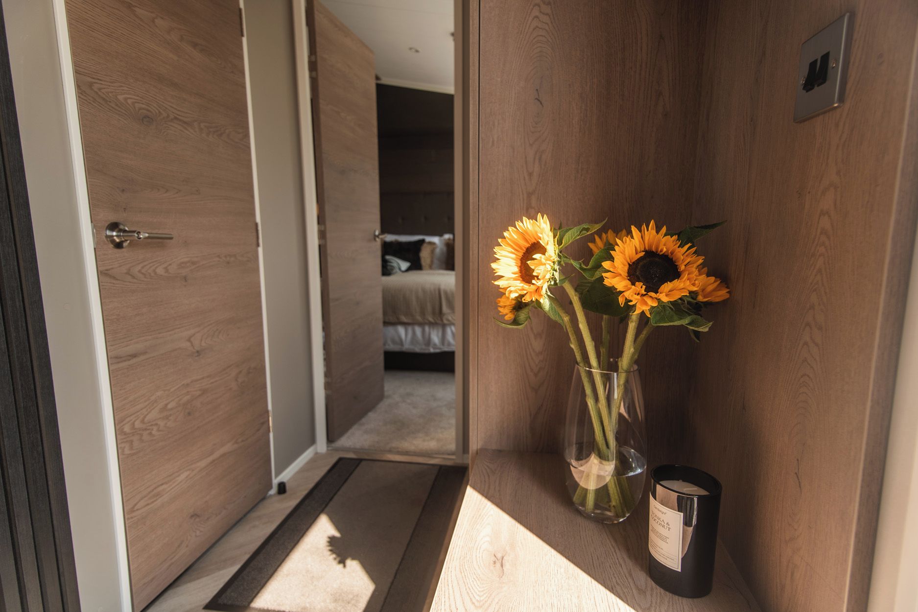Un pasillo con una puerta de madera, una cama al fondo y un florero con girasoles sobre una estantería.