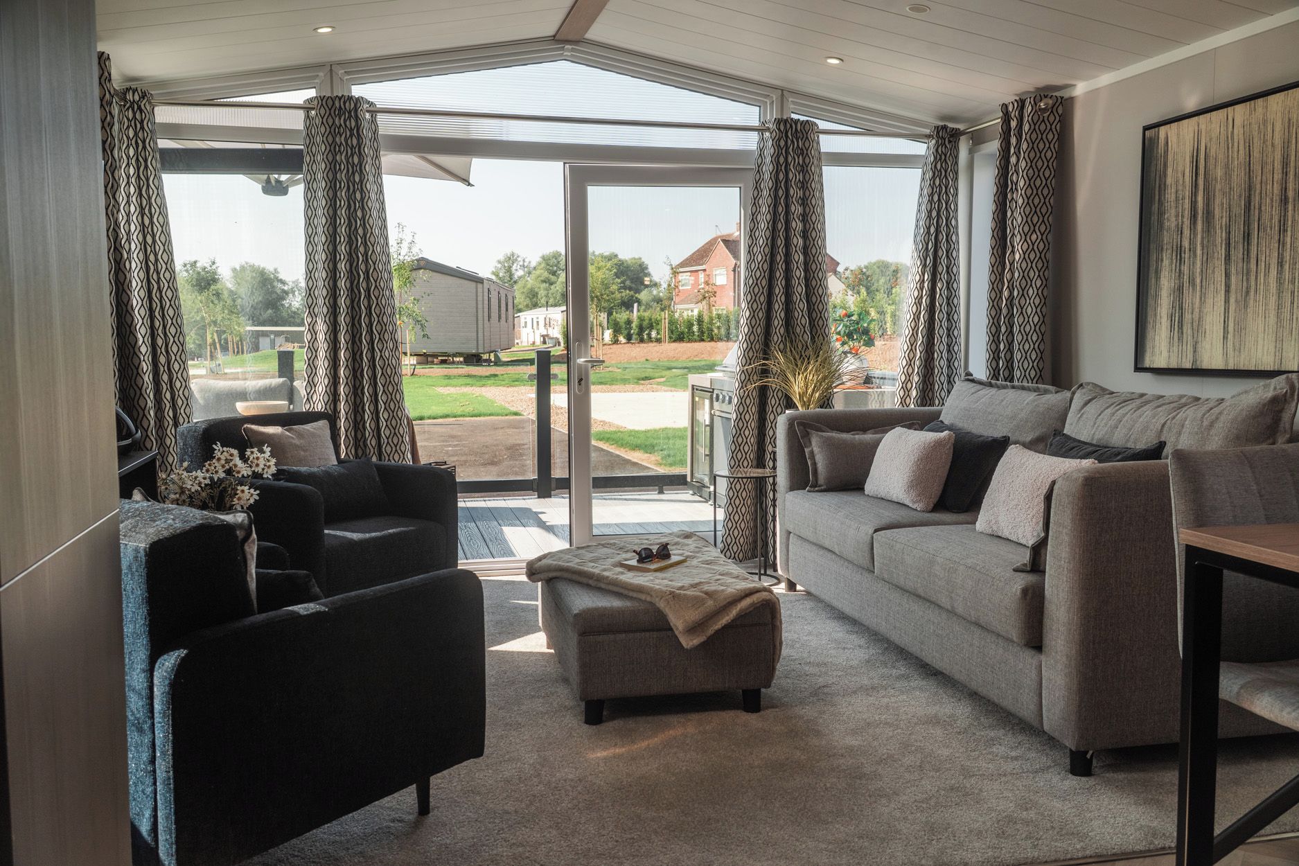 A modern living room with a grey sofa, a black armchair, and a coffee table. There is a large window with patterned curtains, and a view of a residential area outside.