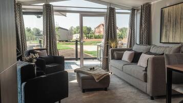 A modern living room with a grey sofa, a black armchair, and a coffee table. There is a large window with patterned curtains, and a view of a residential area outside.