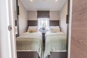 Una habitación estrecha con dos camas, una ventana con cortinas y una pequeña mesa con un florero.