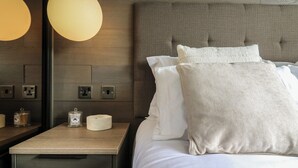 Una cama bien hecha con sábanas y almohadas blancas, una mesita de noche con una lámpara y un atril con una vela y un pequeño recipiente.