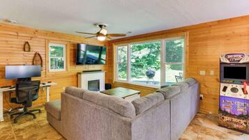 A cozy room with a sofa, a flat-screen TV, a fireplace, and a large window offering a view of greenery.