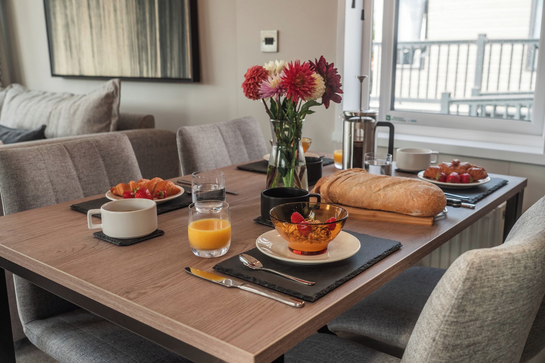 Una mesa de comedor con alimentos para el desayuno, un jarrón con flores y vistas al exterior.