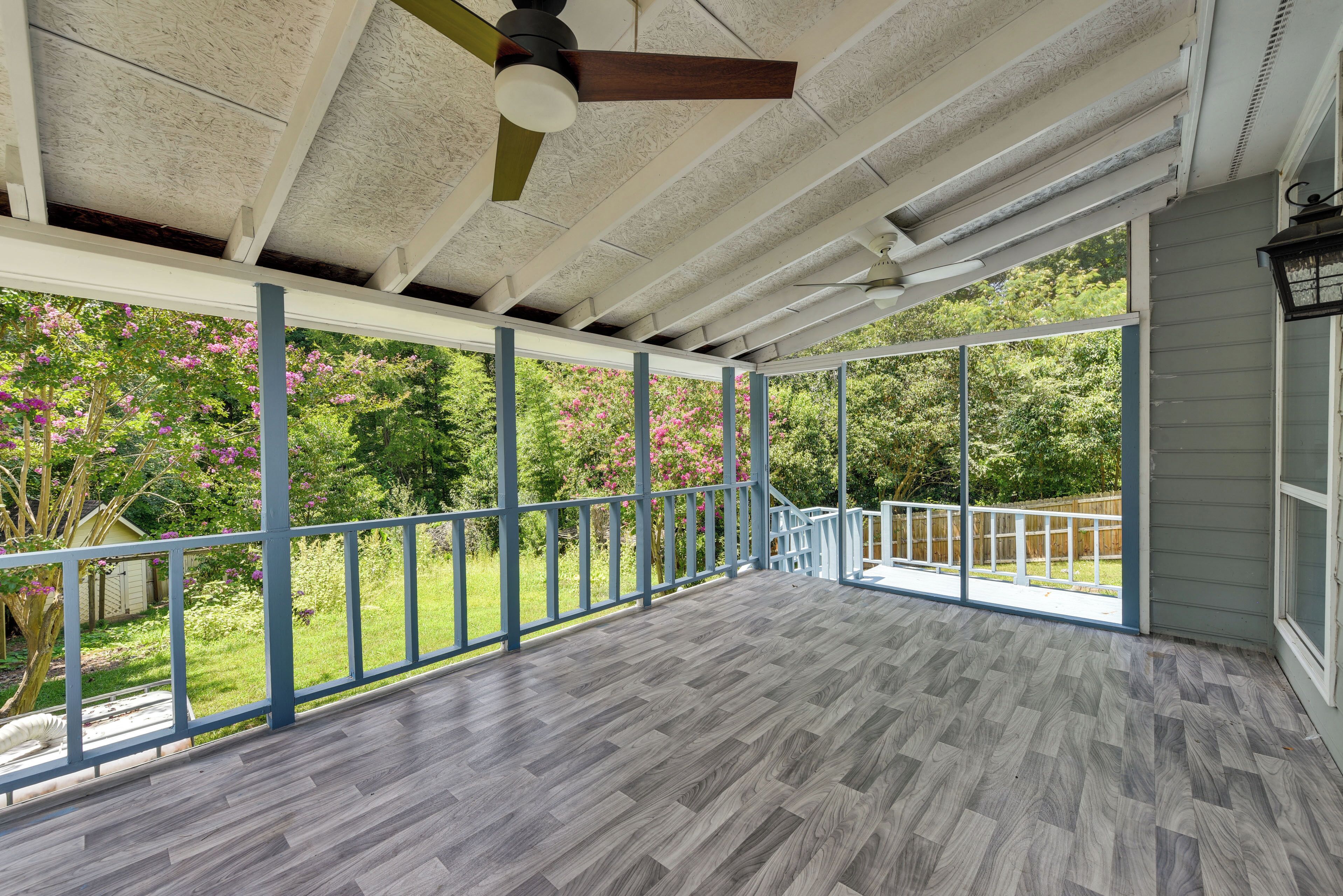 Un espace extérieur abrité, doté d’un plancher en bois, d’un ventilateur de plafond et offrant une vue sur un jardin aux fleurs roses.